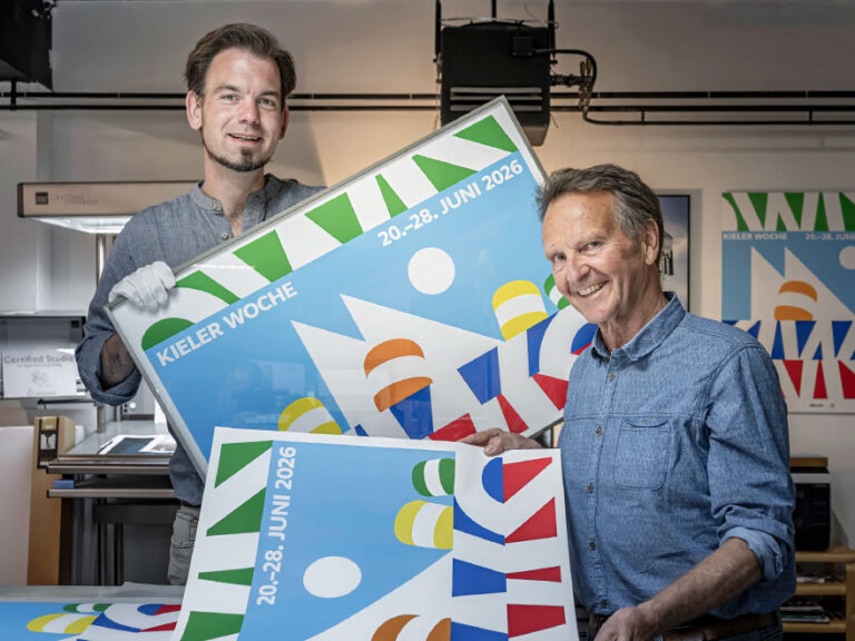 Frank Reimers und Marius Gosch stehen mit einem gerahmten Exemplar des Kieler-Woche-Plakates in der IB-Gosch Werkstatt.