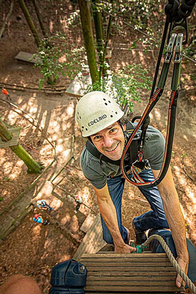 Burkhard Alt klettert gesichert und mit Helm auf eine Plattform im Hochseilgarten. Von oben fotografiert.