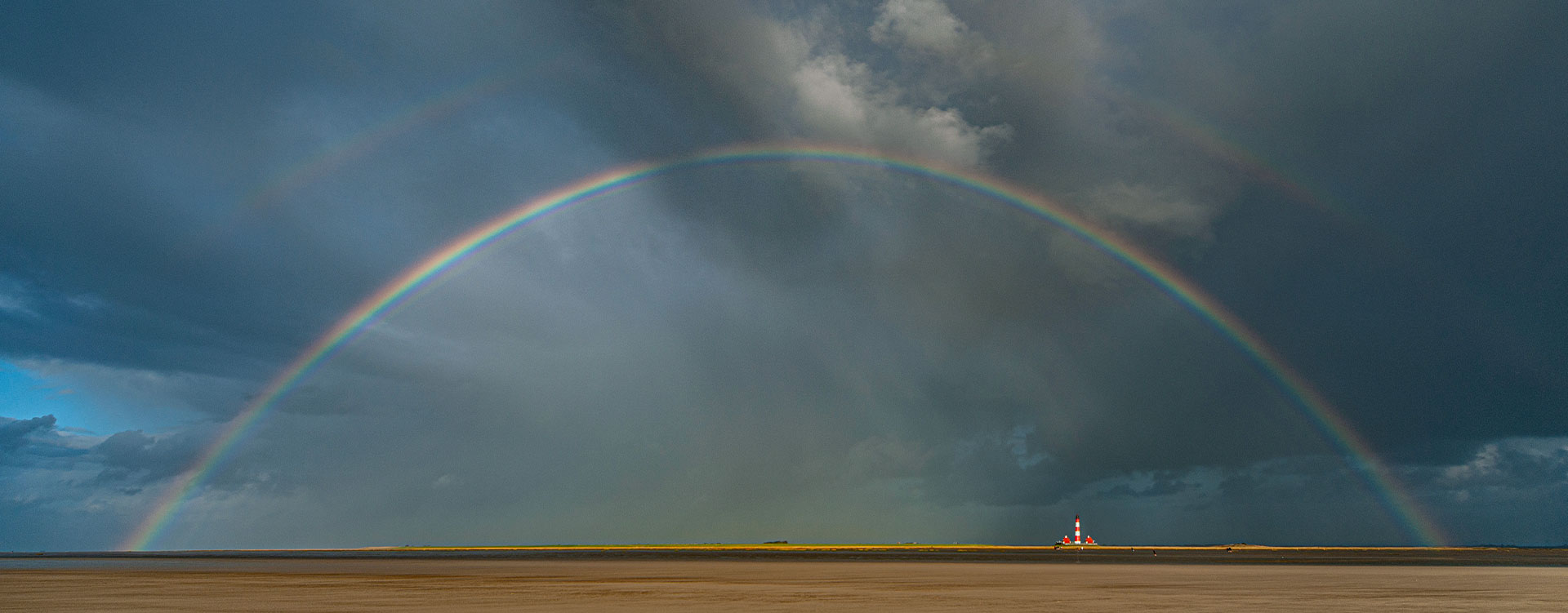 Ein perfekter Regenbogen steht über einem winzig kleinen Leuchtturm in der Ferne. Der Himmel ist atmosphärisch düster und wolkig.